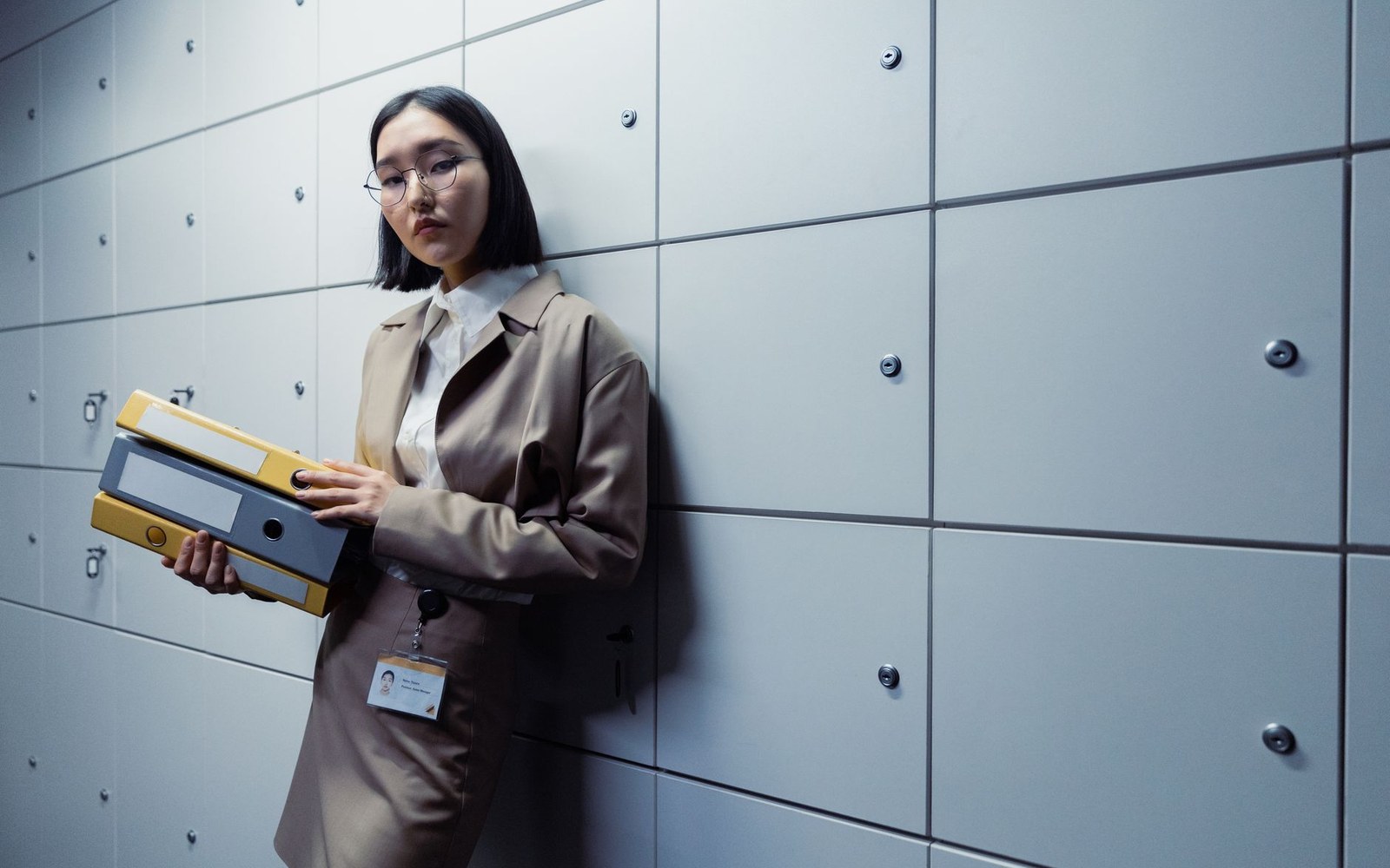 Woman in a trench coat holding binders while leaning against a locker wall