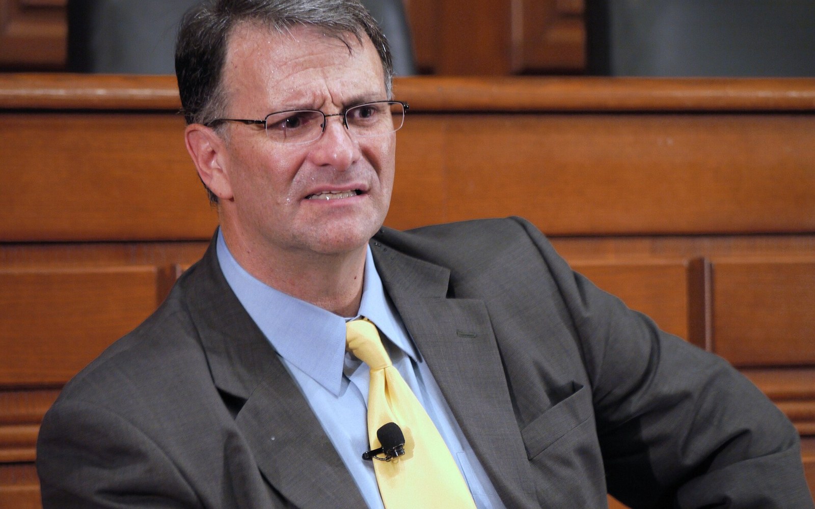 A man in a suit and yellow tie speaks while wearing a clip-on microphone
