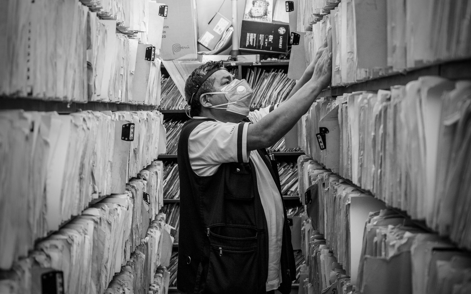 A masked worker reaches for files among crowded shelves stacked with paper records